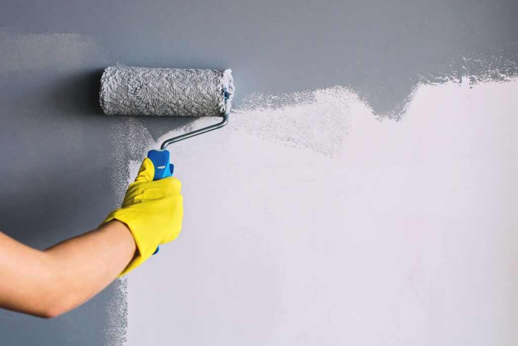 Hand in yellow glove painting wall in gray color with a roller brush- how to apply multiple coats of paint